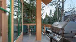 Een terras buiten met een barbecue, houten terras en grote ramen met uitzicht op een bebost gebied.