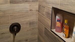 Een douche met een handdouche en bediening aan de wand. Er is een plank met toiletartikelen, waaronder een fles shampoo en een fles lotion.