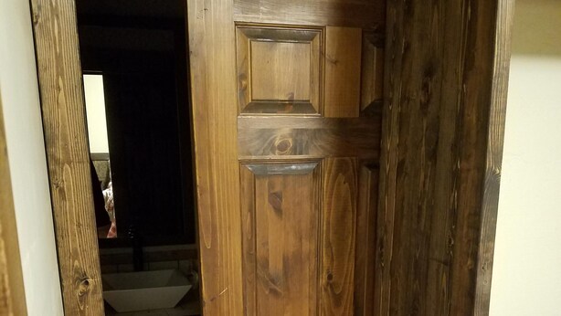 Een houten deur met een sober ontwerp, die leidt naar een ruimte met een zichtbare wastafel en een donker interieur.