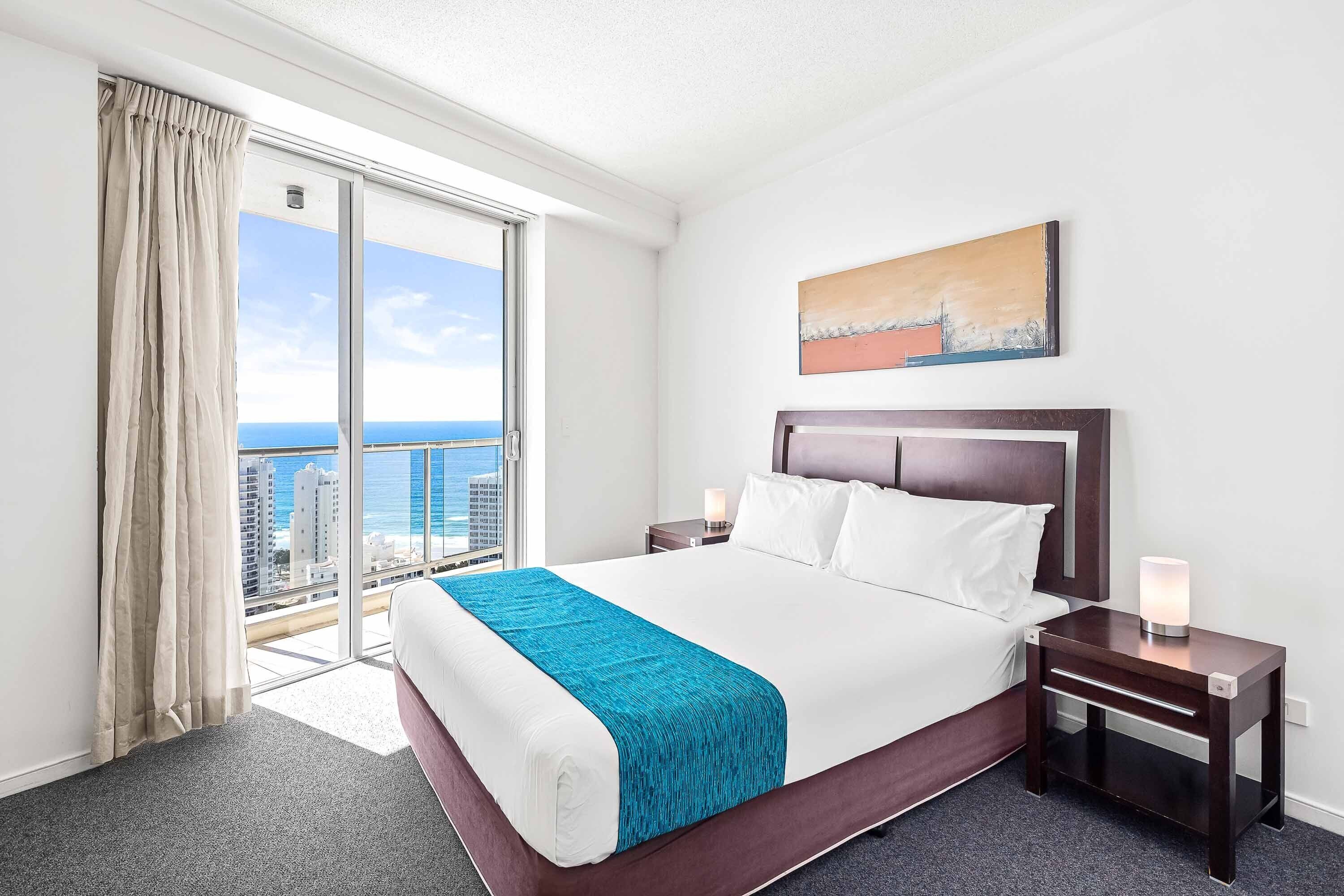 A hotel room with a large bed, a nightstand, a bedside table, a window with curtains, a painting on the wall, and a view of the city and ocean.