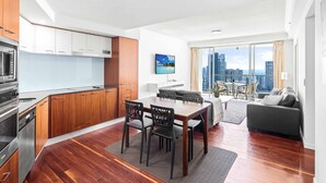 A modern apartment with a kitchen, dining area, and living room. It features wooden flooring, a large window with a city view, and a flat-screen TV mounted on the wall. - Chevron Renaissance Ocean View (Surfers Paradise)