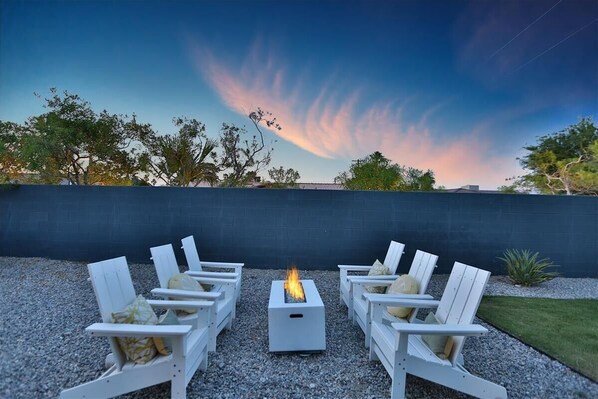 A fire pit with four white Adirondack chairs arranged around it on a gravel surface, set against a dark wall with trees in the background. - Resort Home*The Llama House*Featured on HGTV* Pool and Spa! (Palm Springs)