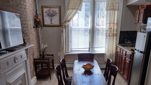 A dining area with a wooden table and chairs, a television on a stand, a potted plant, and a window with curtains. - 2 BEDROOM 3RD FLOOR RENOVATED W/KITCHEN - 3 MINUTES TO SUBWAY (Manhattan)