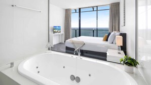 Une chambre d’hôtel moderne dotée d’une grande baignoire, d’un lit offrant une vue sur la ville et d’une petite table de chevet avec une lampe et une plante.