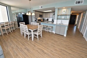 A modern kitchen with a central island, white cabinetry, and a dining area with a wooden table and chairs. - One Ocean Place 605 - Oceanfront in Garden City Beach (Garden City)