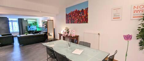 Una habitación de hotel moderna con una mesa de comedor de vidrio, un sofá y un cuadro abstracto grande. Hay dos letreros de 'No Fumar' en la pared.
