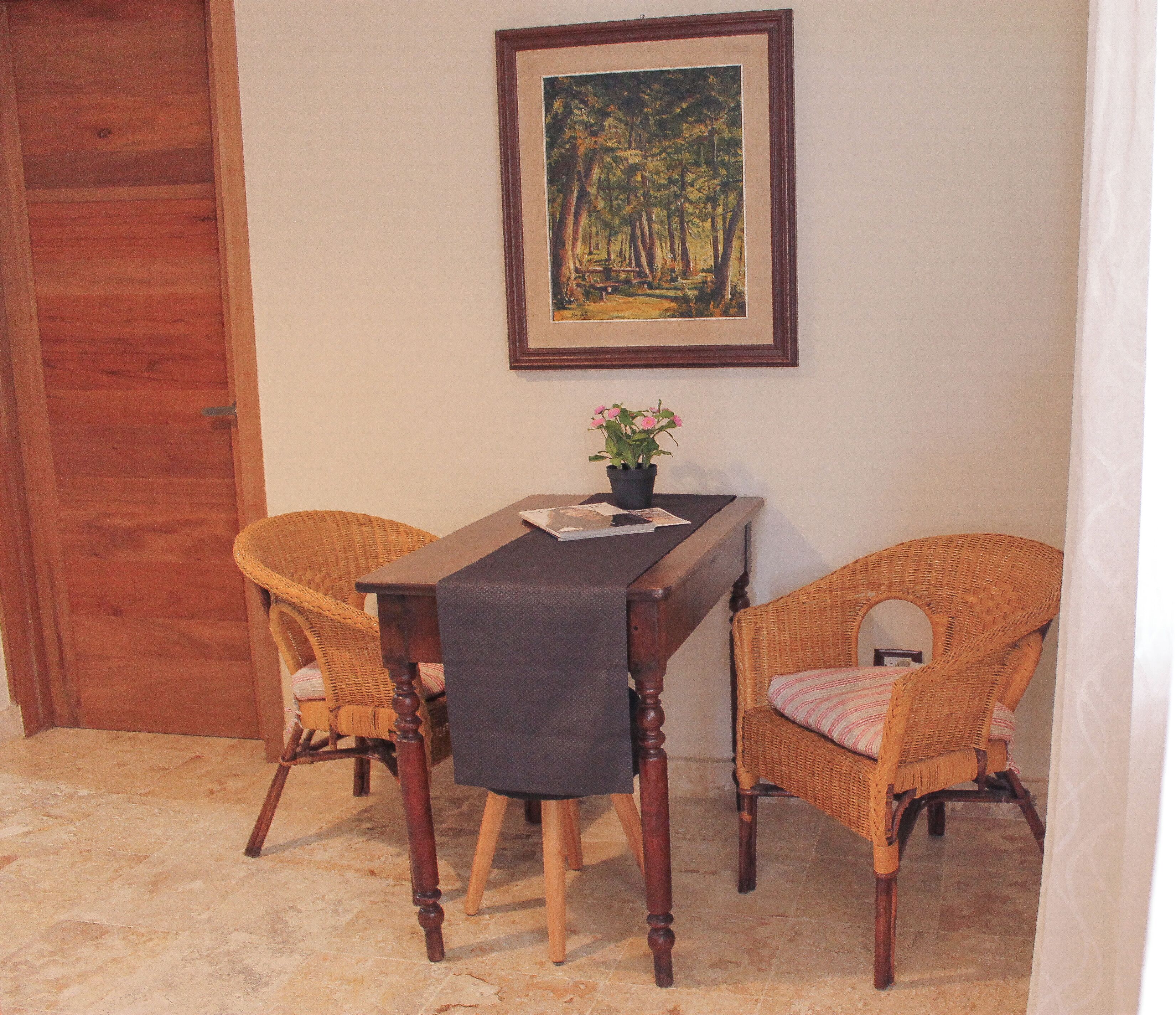 Un comedor con mesa de madera, dos sillas de mimbre, un cuadro en la pared y una planta en maceta.