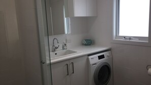 A compact, modern bathroom with a glass shower enclosure, a built-in sink, and a front-loading washing machine. - Gorgeous Stone Cottage on the Seafront (Encounter Bay)