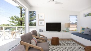 A modern living room with a large TV, comfortable seating, and a balcony with outdoor furniture. - Paddle on Seven Mile (LENNOX HEAD)