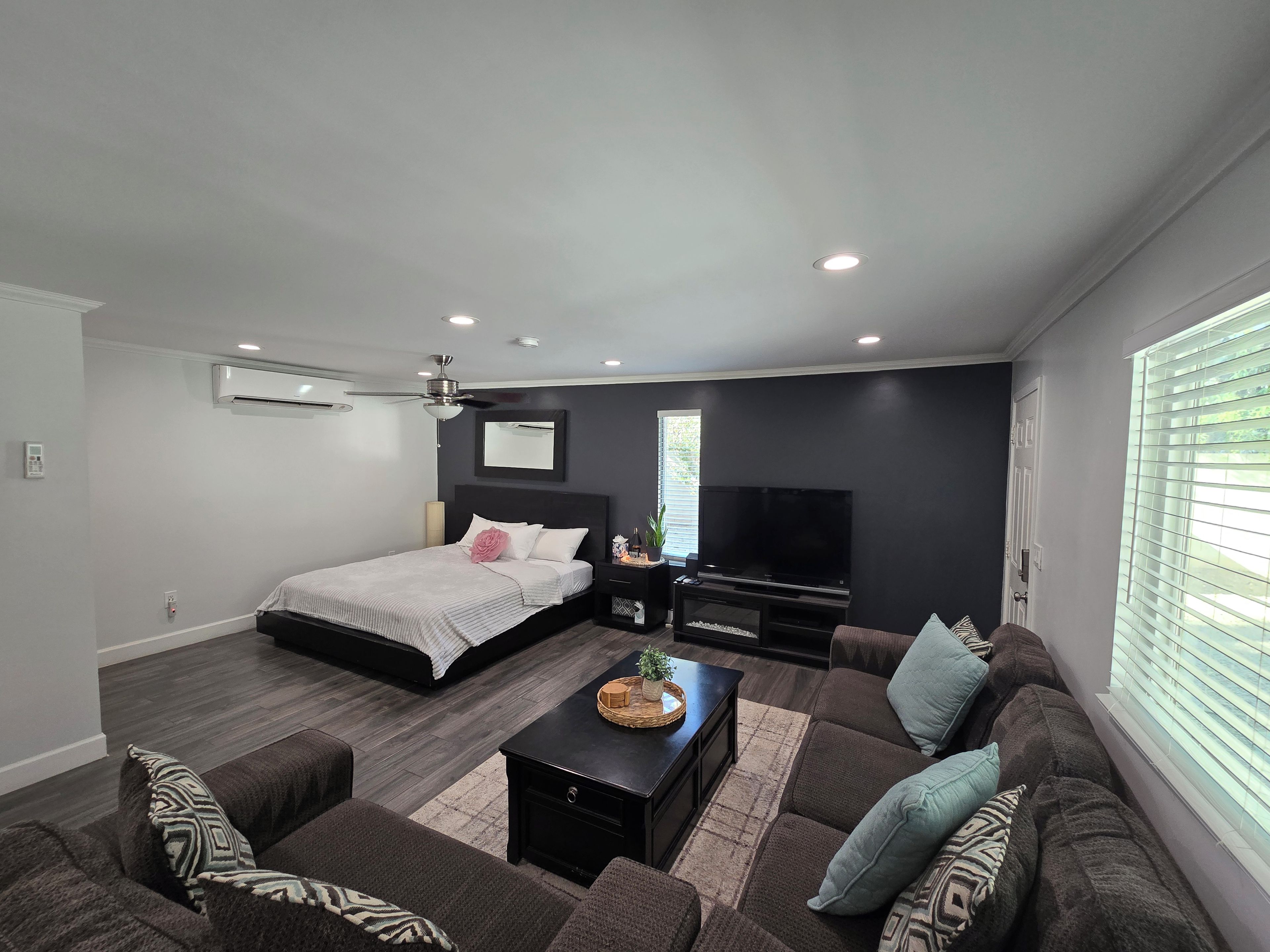 A modern living room with a bed, a flat-screen TV, a ceiling fan, and a large window with blinds.