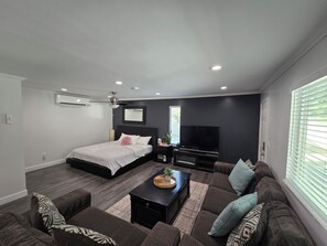 Una sala de estar moderna con una cama, un televisor de pantalla plana, un ventilador de techo y un ventanal con persianas.