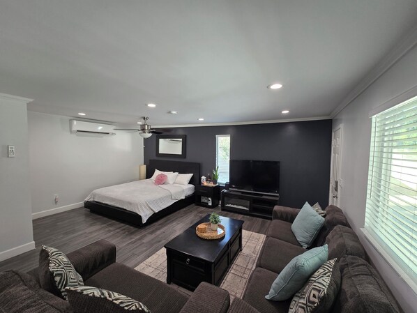 Una sala de estar moderna con una cama, un televisor de pantalla plana, un ventilador de techo y un ventanal con persianas.