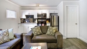A compact living space with a kitchenette, including white cabinets, a microwave, and a refrigerator. There is a sofa with patterned cushions and a coffee table in the foreground.