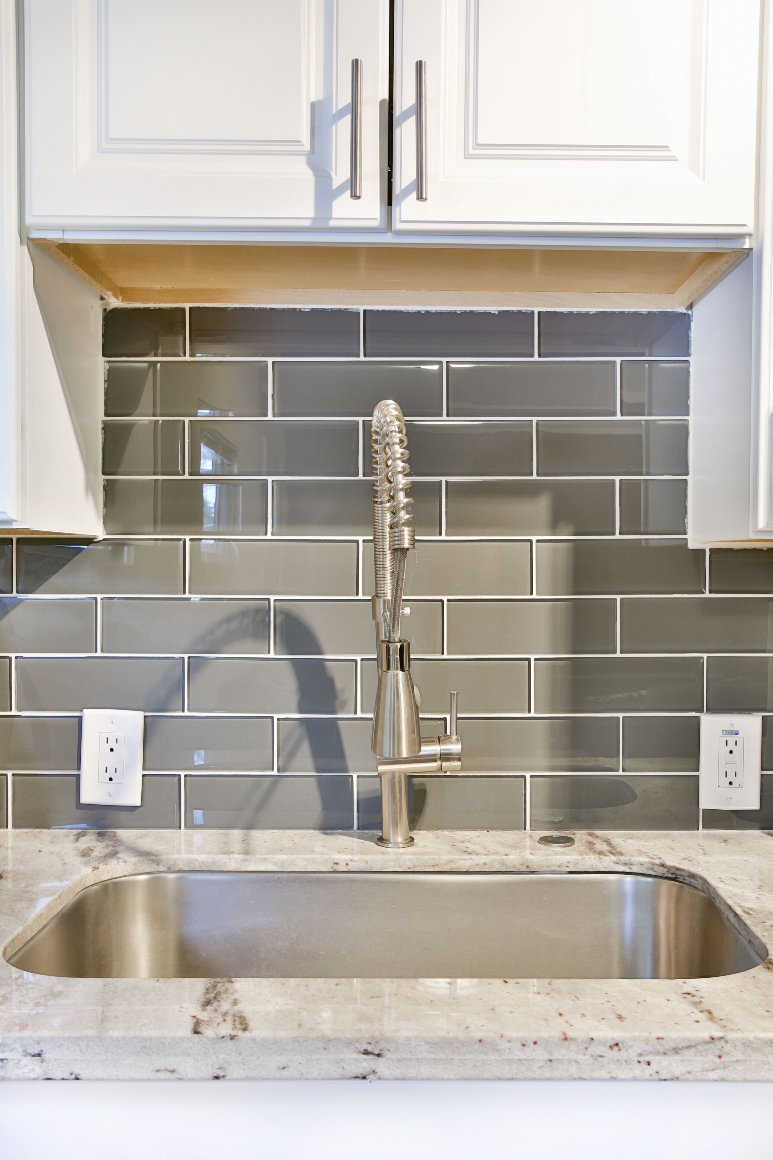 A kitchen with a stainless steel sink, white cabinets, and gray tiled backsplash.