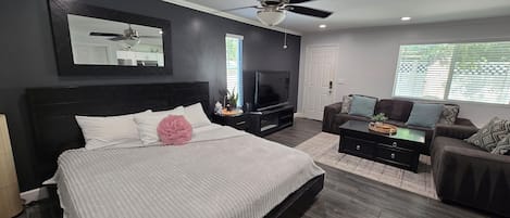 A modern bedroom with a large bed, a ceiling fan, a TV, and a seating area with a sofa and coffee table.