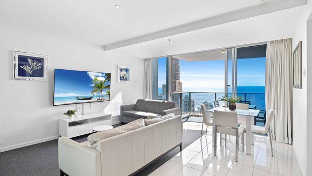 A modern living room with a large flat-screen TV, a grey sofa, a dining table with chairs, and a balcony with a view of the ocean and city skyline. - H Residences Ocean View (Surfers Paradise)