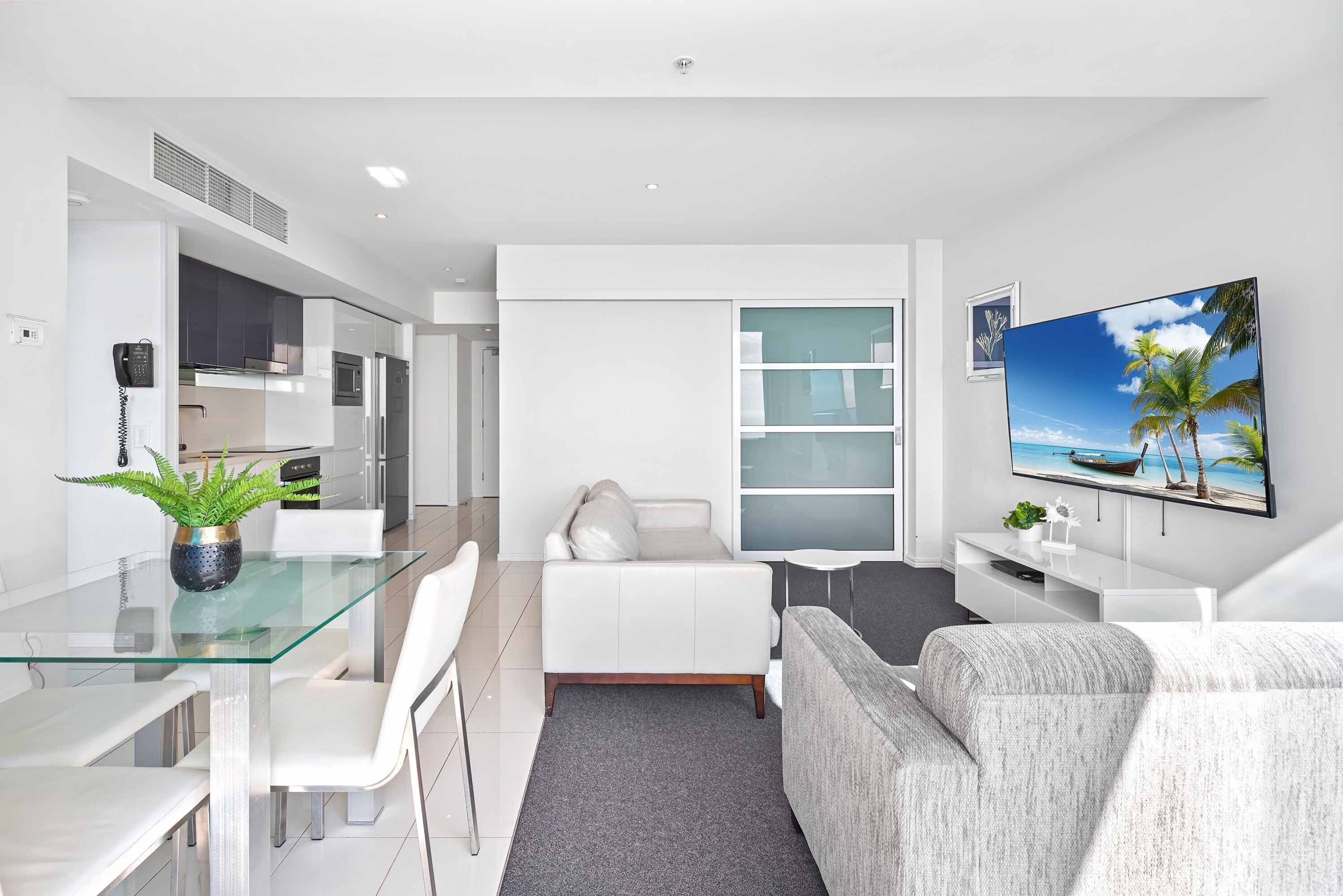 A modern living room with a glass dining table, white sofa, and a flat-screen TV on the wall.
