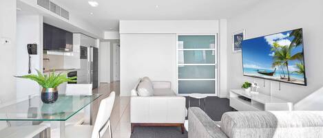 A modern living room with a glass dining table, white sofa, and a flat-screen TV on the wall.