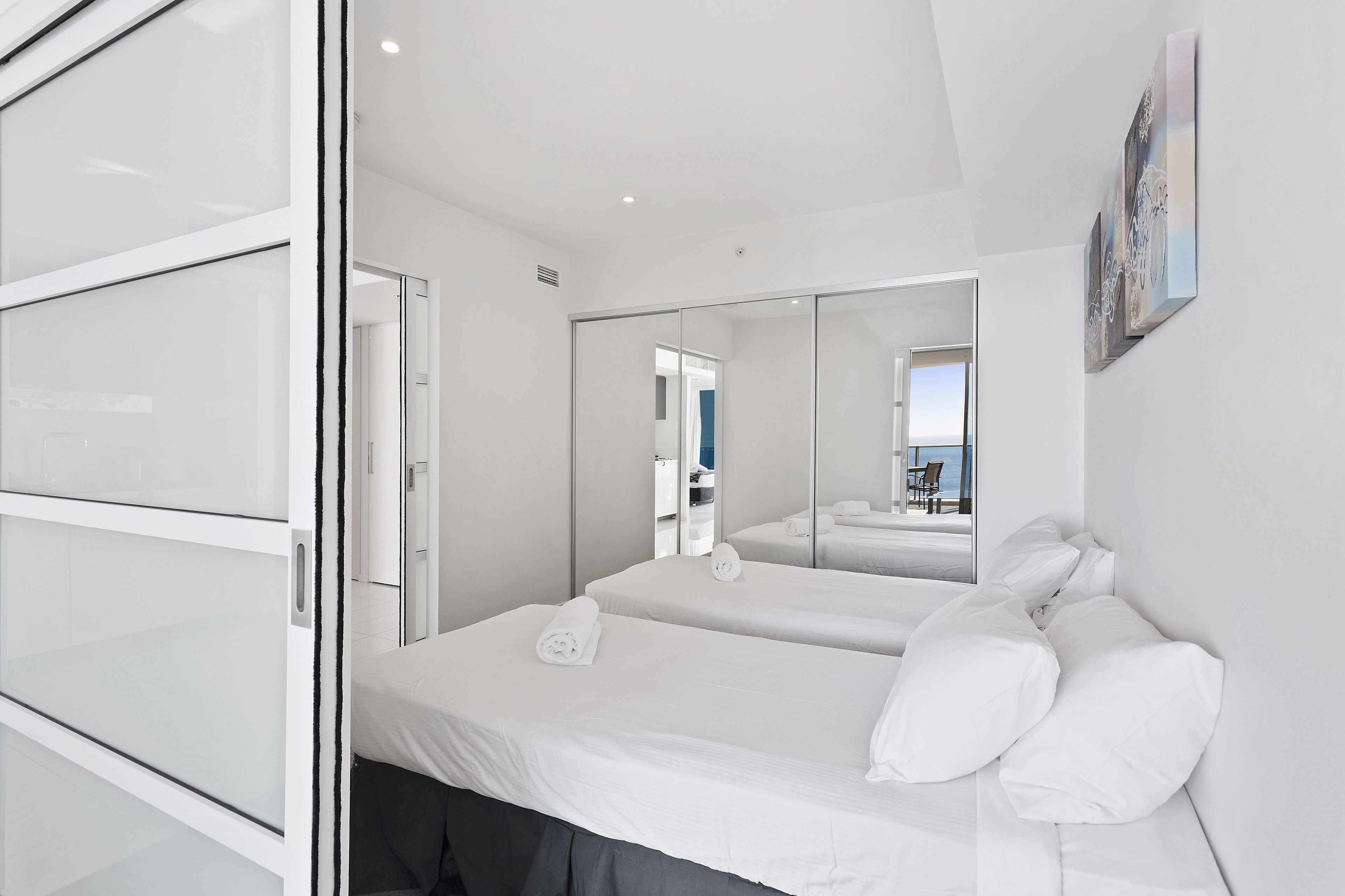 A modern hotel room with a large bed, a glass partition, and a view of the beach.