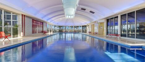 Una amplia piscina interior con paredes de vidrio transparente, sillas rojas y un techo con una lámpara azul.
