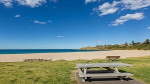 Una playa con mesa de picnic, banco, zona verde y cielo azul despejado.