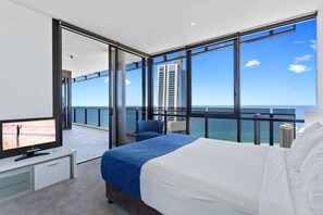 A modern hotel room with a large bed, a TV, and a balcony with a view of the ocean and a tall building.