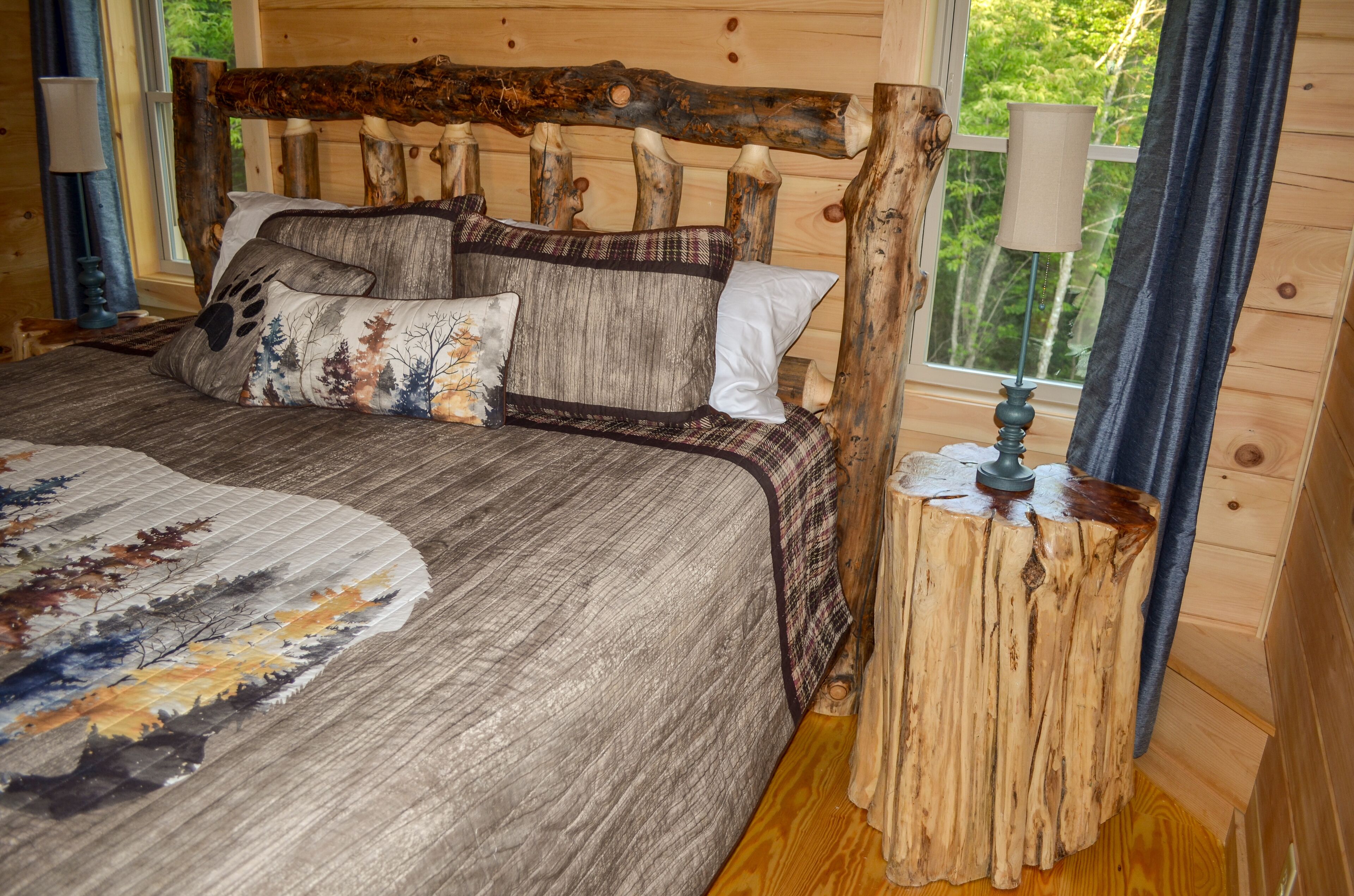 Een bed met een rustiek hoofdbord, een houten nachtkastje met een lamp, en uitzicht op bomen door een raam.