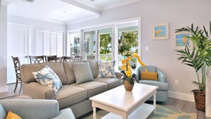 A spacious living room with a large sectional sofa, a coffee table, and a light blue armchair. There is a large window providing a view of the outdoors. - Waterfront 3-Bedroom Condo at Bayfront Suites & Marina- CLEANING FEE INCLUDED (Key West)