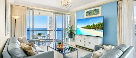 A modern living room with a large TV, comfortable seating, a balcony with a view of the ocean, and a chandelier.