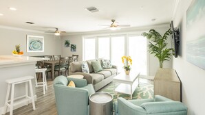 Un salon moderne avec un canapé gris, des fauteuils turquoise et une table basse. On y trouve également un coin repas avec des chaises en bois et une table blanche. Une grande fenêtre laisse entrer la lumière naturelle, et un ventilateur de plafond est visible.