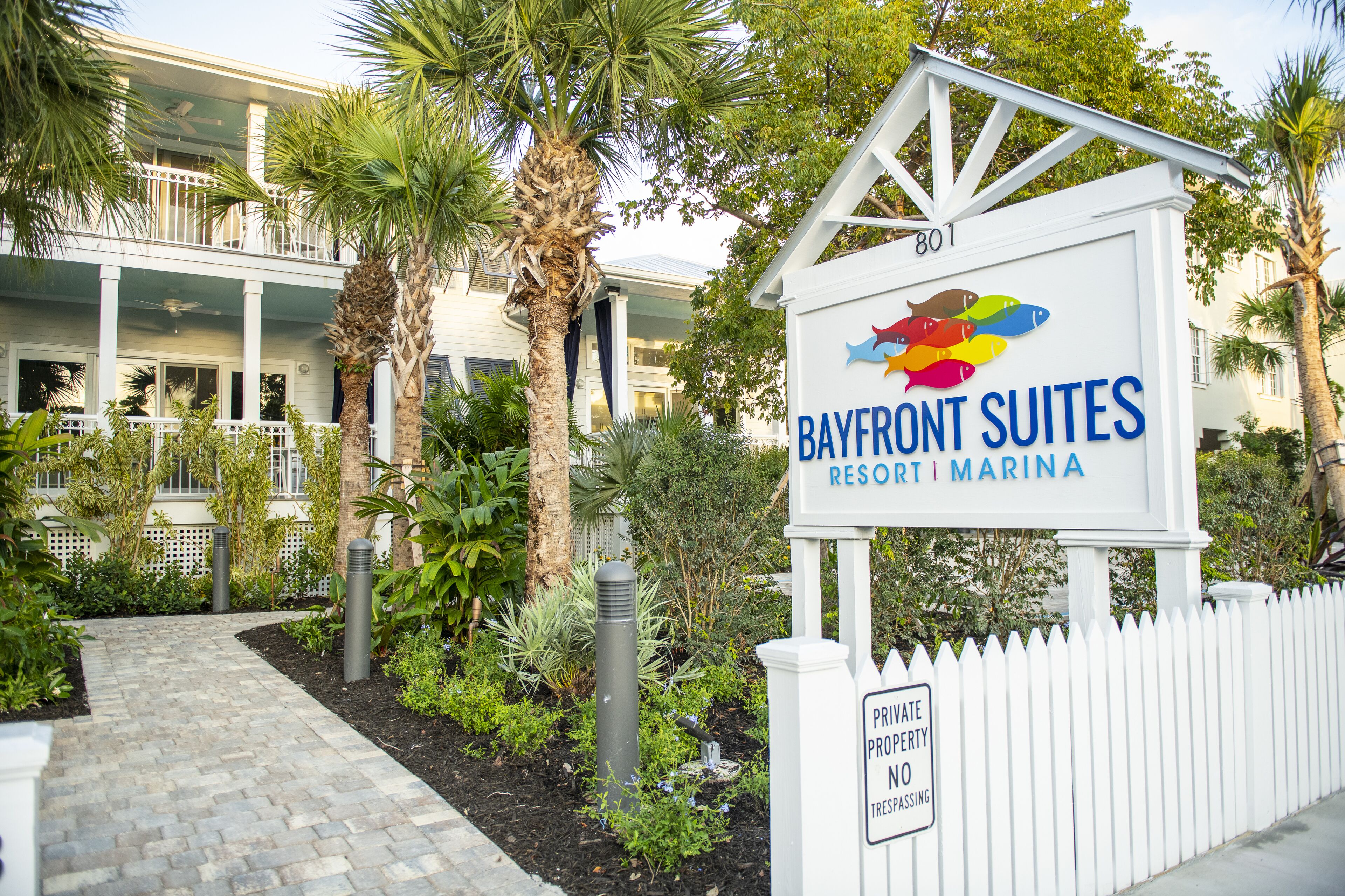 A sign for Bayfront Suites Resort Marina with a white picket fence and tropical landscaping.