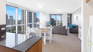 Eine moderne Wohnung mit Küche, Essbereich und Wohnzimmer. Große Fenster bieten Blick auf die Stadt, ein Esstisch mit Stühlen, ein Sofa und eine kleine Küchenzeile mit Arbeitsplatte.