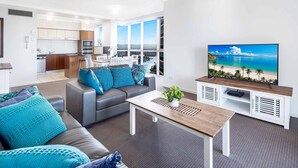 Ein modernes Wohnzimmer mit Sofa, Couchtisch und Fernseher, im Hintergrund eine Küche und ein großes Fenster mit Blick auf den Ozean.