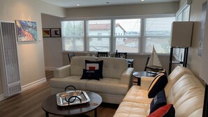 A modern living room with a beige sofa, a coffee table, and a dining area with a small table and chairs. -  Steps to Beach and Bay with Garage Parking (compact car) & Air Conditioning! (Newport Beach)