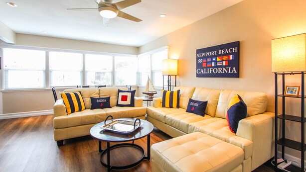 A living room with a sectional sofa, a round coffee table, and a ceiling fan. There is a wall-mounted sign that reads "Newport Beach California." -  Steps to Beach and Bay with Garage Parking (compact car) & Air Conditioning! (Newport Beach)