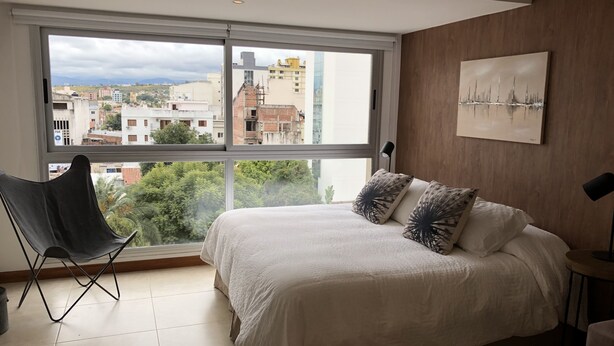 Un dormitorio con un ventanal grande, una cama con ropa blanca, dos almohadones decorativos, una silla negra y una mesita de noche con una lámpara.