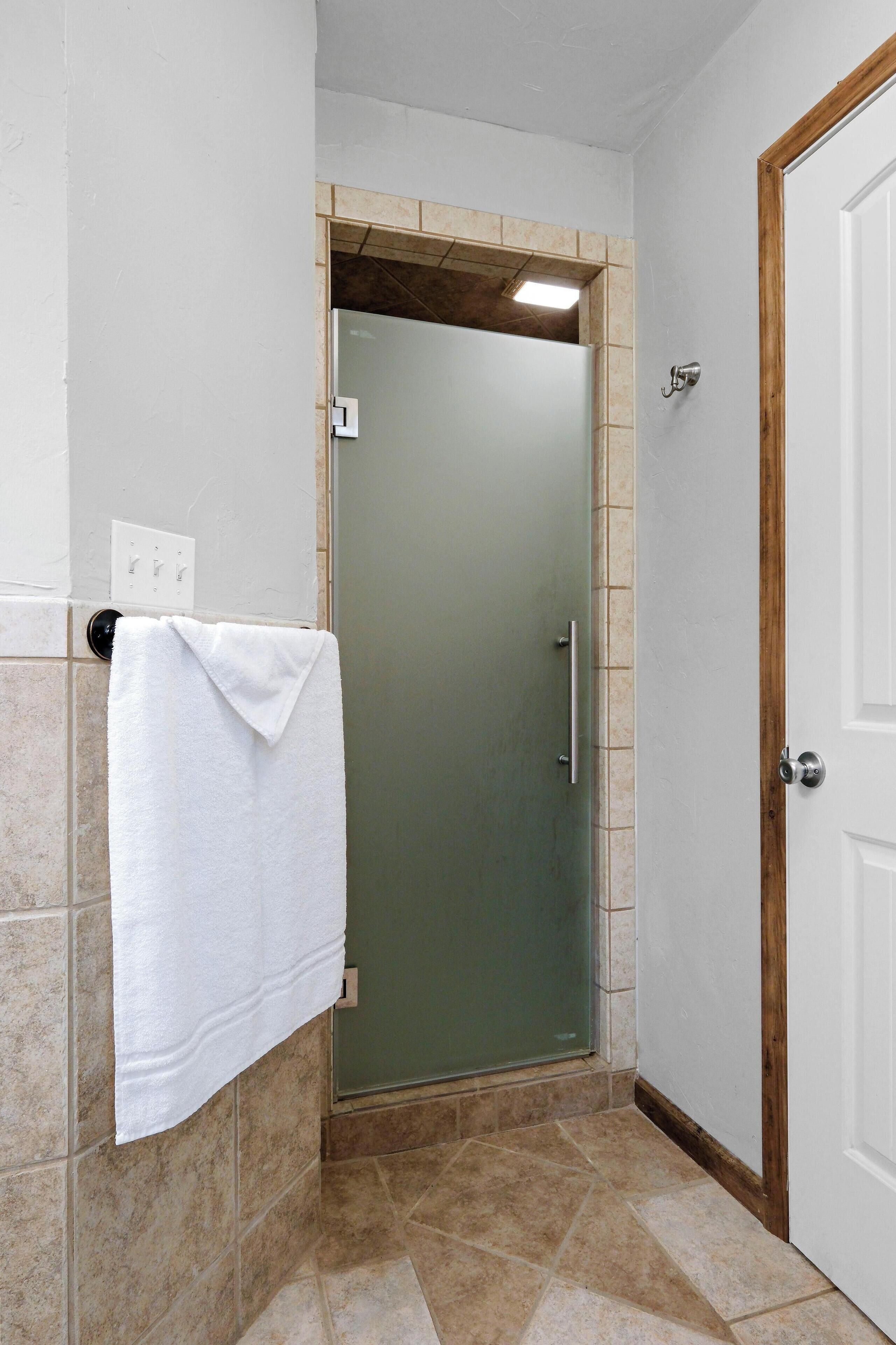 Un baño con cabina de ducha de vidrio, una toalla blanca colgada en la pared y una puerta de madera.