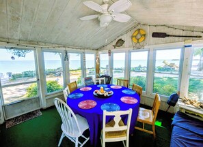 Un área de comedor techada con una mesa dispuesta para cuatro personas, rodeada de amplias ventanas que ofrecen vista al mar y a la playa.