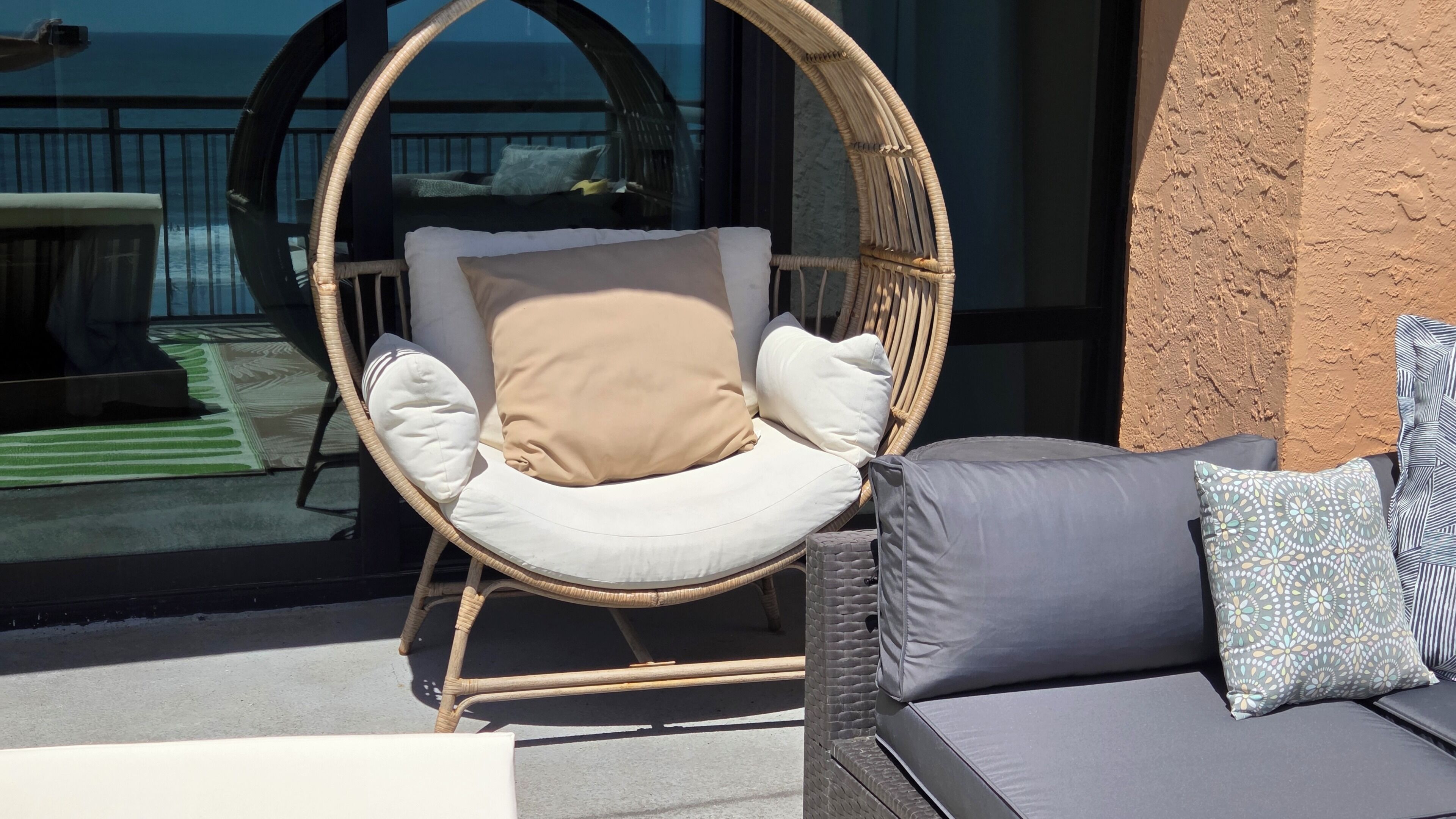 A wicker chair with cushions, a sofa with patterned pillows, and a view of the sea through a glass door.