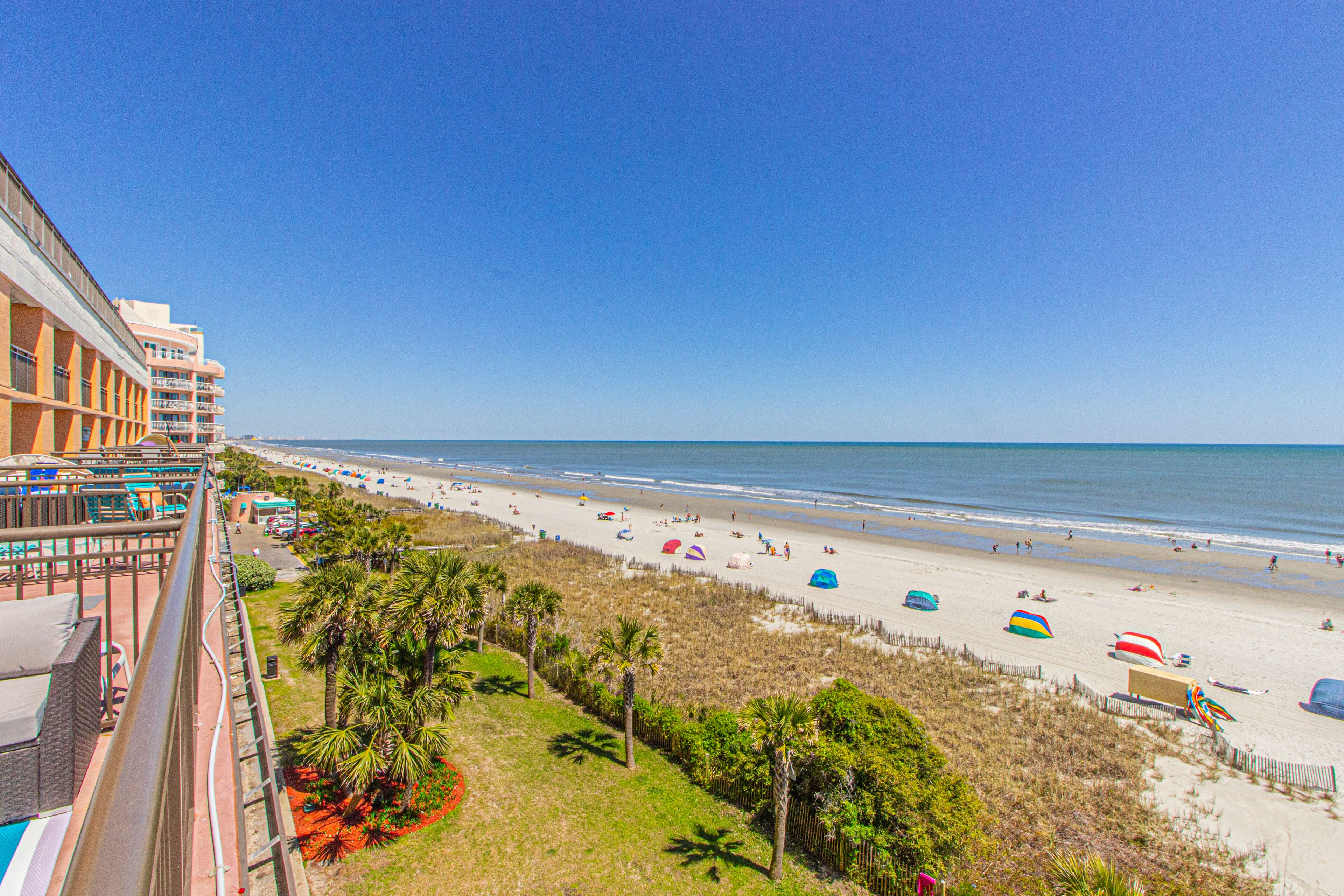 Blick auf den Strand vom Hotelbalkon mit blauem Himmel, Palmen und Strandsonnenschirmen.