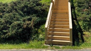 Eine hölzerne Treppe führt einen grasbewachsenen Hügel hinauf, im Hintergrund ein Haus.