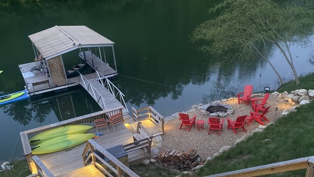 Eine Holzstege führt in einen See, am Ufer steht ein kleines Gebäude und rote Stühle.