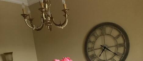 A dining table with white chairs, a vase with flowers, a clock, and a chandelier.