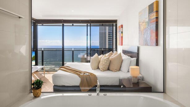 A modern bedroom with a large bed, a bedside table with a lamp, a view of the ocean, and a balcony.