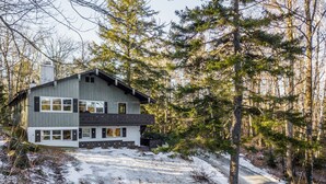 A modern two-story house with a sloped roof, large windows, and a stone foundation, surrounded by a forested area with snow on the ground. - Killington Chalet - Great Space and Location! (Killington)
