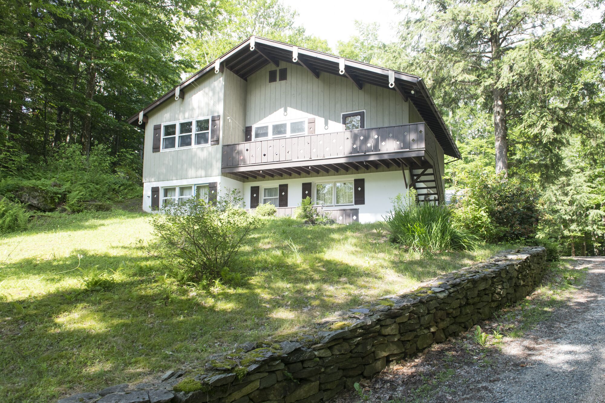 Une cabane en bois à deux étages, dotée d’un balcon, entourée d’une végétation luxuriante et d’un mur de pierre.