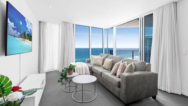 A modern living room with a grey sofa, white coffee table, and a TV on the wall. It features a large window with a view of the ocean and a building in the distance. - H Residences Ocean View (Surfers Paradise)