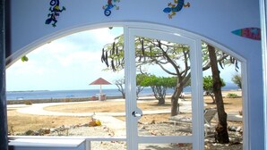 A view from a room with a glass door leading to an outdoor area with a sandy path, trees, and a beach in the background. - Spacious Oceanfront Studio Condo A3 at Sand Dollar Condominiums (Kralendijak)