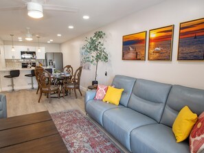 A modern living room with a blue sofa, a dining area with wooden chairs, and a kitchen in the background.