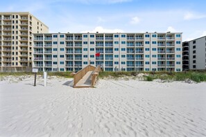 Et hotel ved stranden med flere altaner, en sandstrand foran og en træpromenade, der fører til indgangen.